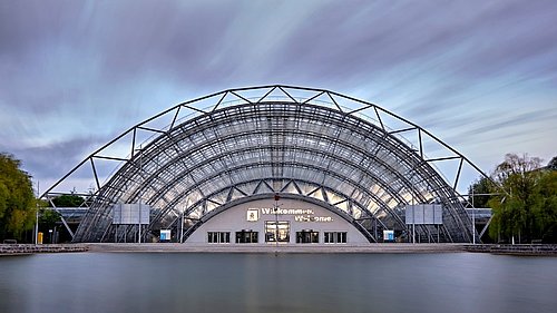 Foto, modernes Gebäude mit halbkreisförmiger Stahl-Glas-Fassade am Wasser, darüber bewölkter Himmel mit Lichtstreifen.
