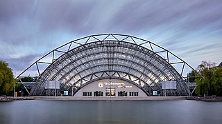 Foto, modernes Gebäude mit halbkreisförmiger Stahl-Glas-Fassade am Wasser, darüber bewölkter Himmel mit Lichtstreifen.