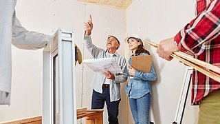 Foto, vier Personen in einem Raum auf einer Baustelle. Im Vordergrund hält eine Person ein Fensterbauteil, eine weitere Person eine Wasserwaage. Im Hintergrund studieren ein Mann und eine Frau einen Baupläne, der Mann zeigt auf etwas außerhalb des Bildauschnittes.