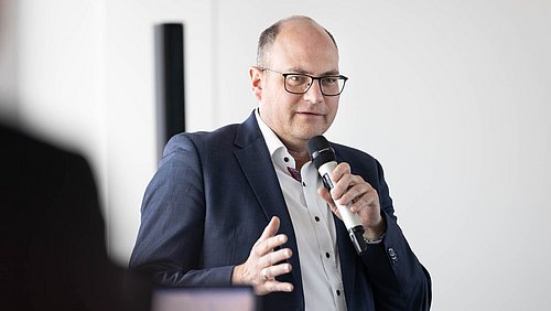 Foto, Mann in Anzug mit Mikrofon in der rechten Hand gestikulierend während einer Rede.