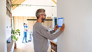 Foto, Mann steht an einer Wand und bedient ein Wandthermostat in einem hellen, modernen Raum mit sichtbarem Dachstuhl und einer weiteren Person im Hintergrund.
