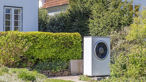 Foto, eine weiße Wärmepumpe steht neben einem gepflegten Garten mit grünen Sträuchern und einem gepflasterten Weg. Im Hintergrund sind Wohnhäuser sichtbar.