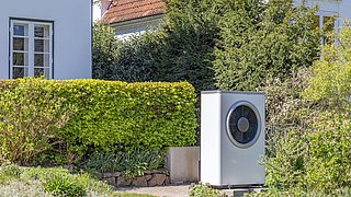 Foto, eine weiße Wärmepumpe steht neben einem gepflegten Garten mit grünen Sträuchern und einem gepflasterten Weg. Im Hintergrund sind Wohnhäuser sichtbar.