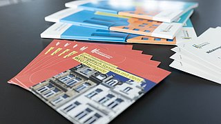 Foto, mehrere Informationsbroschüren liegen auf einem schwarzen Tisch, die vorderste Broschüre zeigt ein Gebäude.