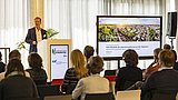 Foto, ein Redner hält einen Vortrag auf einer Konferenz, wobei auf einem großen Bildschirm eine Präsentation über Beispiele und Konzepte für die Sanierung von Quartieren gezeigt wird.