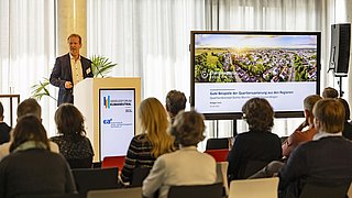 Foto, ein Redner hält einen Vortrag auf einer Konferenz, wobei auf einem großen Bildschirm eine Präsentation über Beispiele und Konzepte für die Sanierung von Quartieren gezeigt wird.