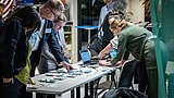 Foto, mehrere Personen stehen an einem Tisch mit ausgebreiteten Flyern und Broschüren, eine Person arbeitet an einem Laptop.