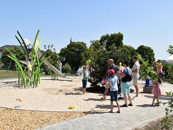 Foto, Kinder und Erwachsene spielen und beobachten auf einem Spielplatz mit Klettergerüst und Rutsche im Freien bei sonnigem Wetter