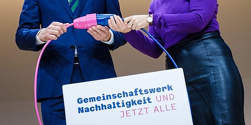 Foto, zwei Personen stecken symbolisch zwei Stecker zusammen, an denen das Logo des Gemeinschaftswerks Nachhaltigkeit befestigt ist.