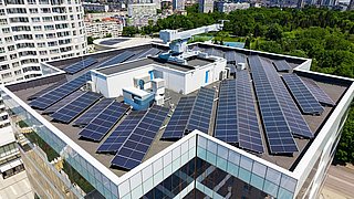 Foto, Dach eines modernen Gebäudes mit zahlreichen Solarpaneelen und umliegender Stadtlandschaft.