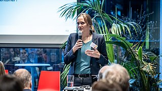 Foto, Frau mit Mikrofon und Namensschild steht vor Publikum, im Hintergrund große grüne Pflanze und Projektionsfläche mit Text.