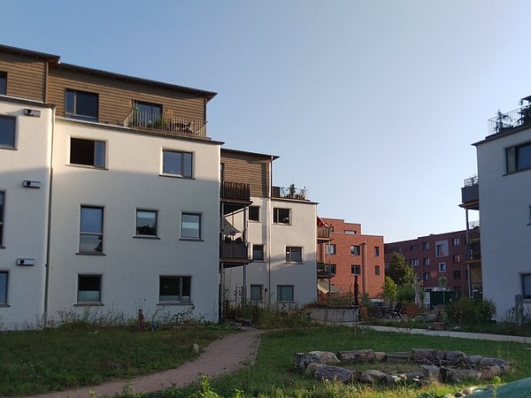 Foto, Mehrgeschossige Wohnhäuser mit weißer Fassade und dunklen Fensterrahmen stehen auf einer Grünfläche. Die Gebäude haben Balkone mit Pflanzen und Holzelementen im oberen Bereich. Im Vordergrund verläuft ein schmaler Weg durch das Gras, daneben liegt eine kreisförmige Steinfläche. Im Hintergrund sind weitere Wohnhäuser in rötlichen Farbtönen sichtbar. Der Himmel ist klar und blau.