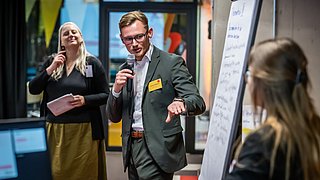 Foto, Mann im Anzug hält Mikrofon und zeigt auf Flipchart mit Notizen, zwei Frauen hören zu, bunte Raumgestaltung im Hintergrund.