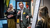 Foto, Mann im Anzug hält Mikrofon und zeigt auf Flipchart mit Notizen, zwei Frauen hören zu, bunte Raumgestaltung im Hintergrund.