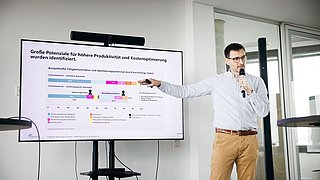 Foto, Mann hält Mikrofon und zeigt auf Bildschirm mit Präsentation zu Produktivität und Kostenoptimierung in Büro mit Glasfenstern