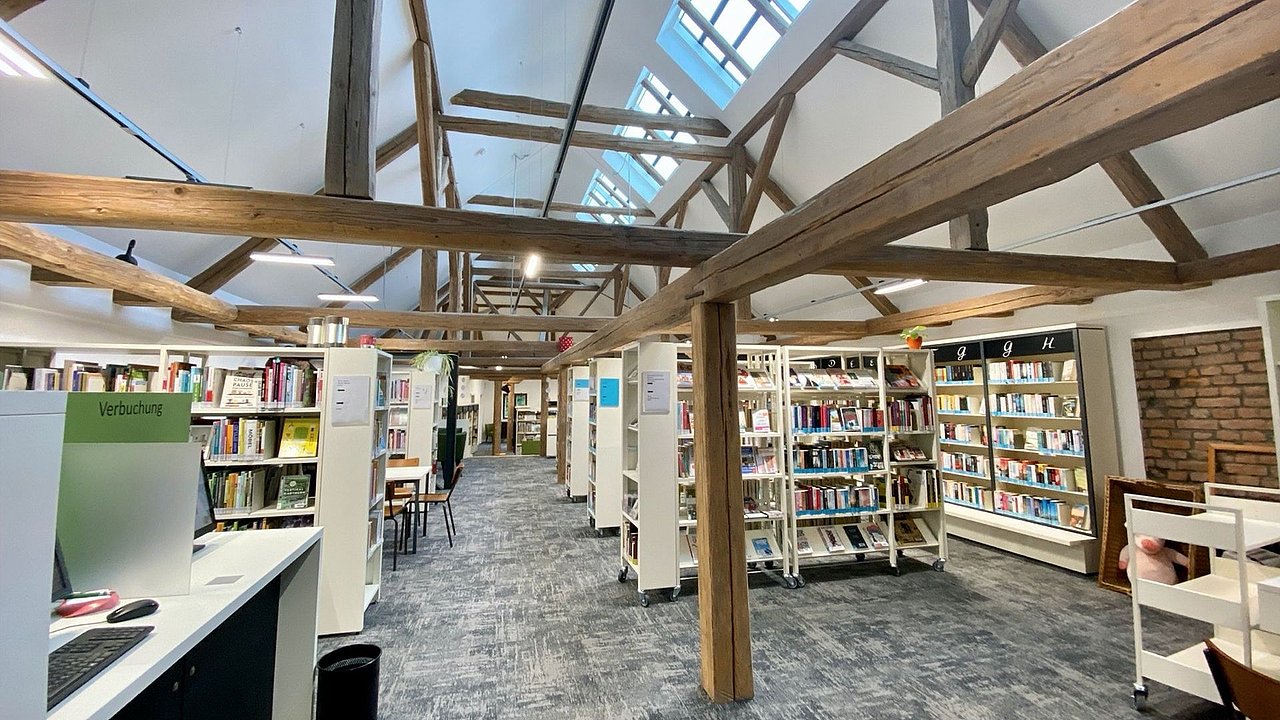 Foto, Innenraum einer Bibliothek mit Holzträgern, Regalen voller Bücher und einem Arbeitsplatz mit Computer