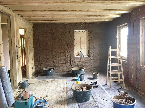 Foto, Innenraum mit teilweise verputzter Lehmwand, Leiter, Eimern und Baumaterialien auf Holzboden