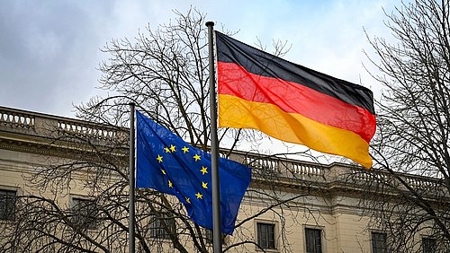 Foto, die wehenden Flaggen der Europäischen Union und Deutschlands vor einem historischen Gebäude und kahlen Bäumen.