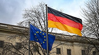 Foto, die wehenden Flaggen der Europäischen Union und Deutschlands vor einem historischen Gebäude und kahlen Bäumen.