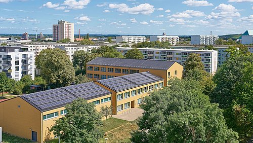 Foto, mehrere Wohngebäude in einer Stadt, im Vordergrund ein gelbes Schulgebäude mit Solarpaneelen auf dem Dach, umgeben von Bäumen.