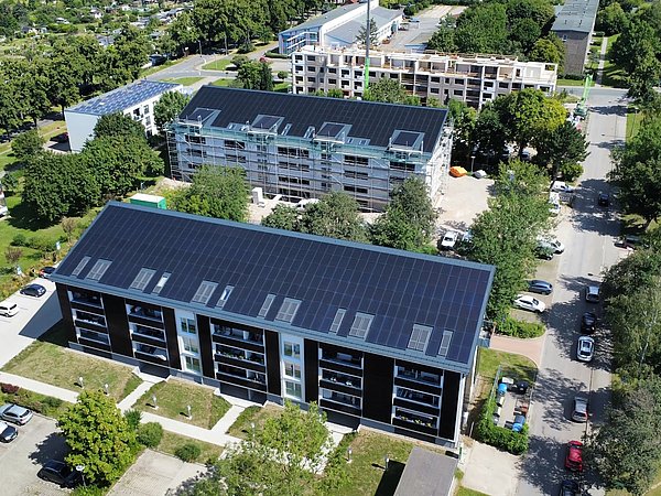 Luftaufnahme von mehreren Wohngebäuden mit Solarpaneelen auf den Dächern und umgeben von Bäumen und Straßen.