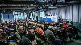 Foto, mehrere Personen sitzen in einem Saal und hören einem Podium mit vier Sprechern zu, die vor einer Leinwand mit Präsentationstext sitzen.