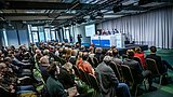 Foto, mehrere Personen sitzen in einem Saal und hören einem Podium mit vier Sprechern zu, die vor einer Leinwand mit Präsentationstext sitzen.