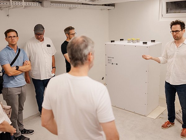 Mann in weißem Hemd und Jeans spricht vor einer Gruppe von Menschen in einem Heizungskeller mit weißer Wand und Fenster vor einem Roots Hub.