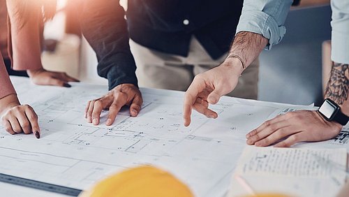 Foto, mehrere Personen zeigen mit den Händen auf einen großen Bauplan auf einem Tisch.