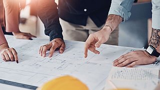 Foto, mehrere Personen zeigen mit den Händen auf einen großen Bauplan auf einem Tisch.