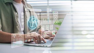 Foto, Person tippt auf Laptop-Tastatur vor halb geschlossenen Jalousien mit eingeblendeten transparenten Smart-Home-Symbolen.