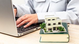 Foto, Modell eines modernen Hauses mit Garten auf einem Buch vor einer Person, die an einem Laptop tippt.