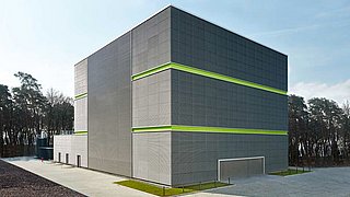 Foto, Großes graues Gebäude mit zwei grünen horizontalen Streifen, umgeben von Wald und gepflastertem Hof