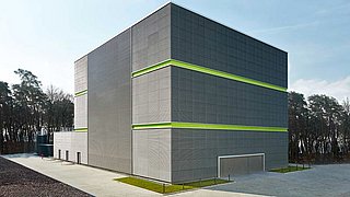 Foto, Großes graues Gebäude mit zwei grünen horizontalen Streifen, umgeben von Wald und gepflastertem Hof