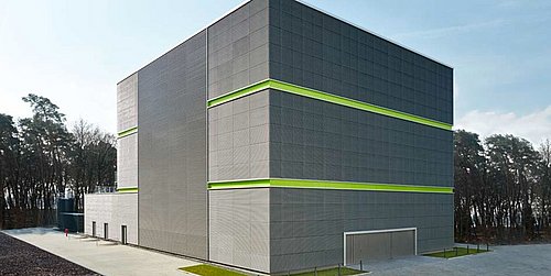 Foto, Großes graues Gebäude mit zwei grünen horizontalen Streifen, umgeben von Wald und gepflastertem Hof