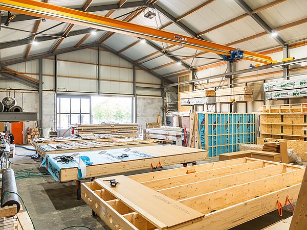 Foto, eine helle Werkshalle mit Holzkonstruktionen, Kränen, Arbeitsmaterialien und gestapelten Holzplatten.