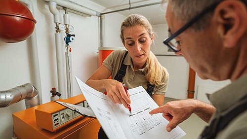 Foto, eine Monteurin und ein Monteur stehen in einem Heizungskeller und schauen gemeinsam auf ein technisches Dokument.