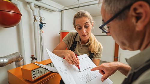 Foto, eine Monteurin und ein Monteur stehen in einem Heizungskeller und schauen gemeinsam auf ein technisches Dokument.