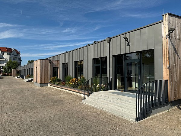 Foto, Modernes, langgestrecktes Gebäude mit grauer Fassade und Holzverkleidung, Treppe mit schwarzem Geländer, blauer Himmel