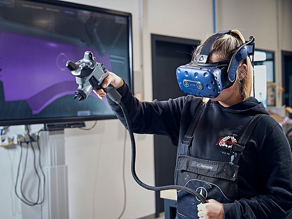 Foto, eine Person trägt eine Virtual-Reality-Brill und hält in einer gehobenen Hand eine Spritzpistole. Im Hintergrund ist ein Bildschirm an der Wand angebracht. 