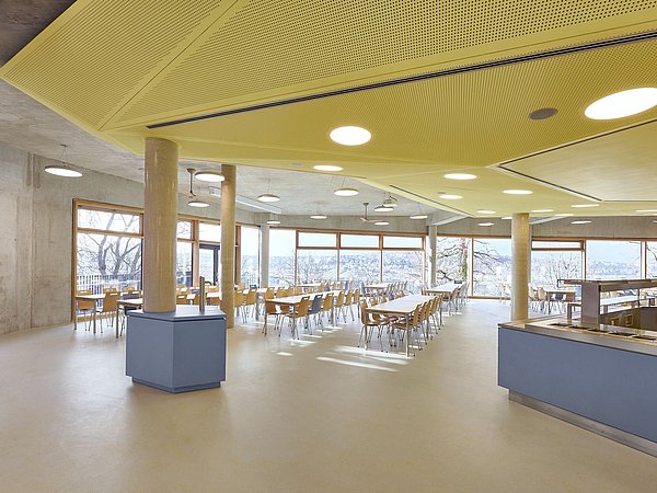 Foto, Großer, heller Speisesaal mit gelber Decke, vielen Holztischen und Stühlen, großen Fenstern und Blick auf Bäume