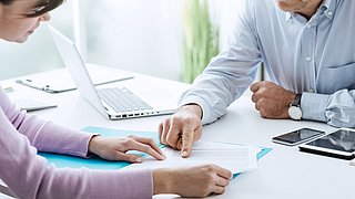 Foto, ein Mann und eine Frau sitzen sich am Schreibtisch gegenüber und schauen gemeinsam auf ein Dokument.