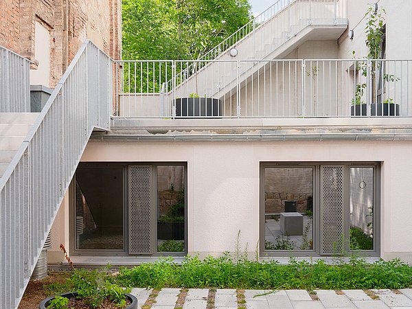 Foto, Innenhof mit moderner, weißer Fassade, großen Fenstern, Metallgeländern und einem runden Pflanzkübel auf gepflastertem Boden.