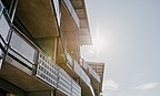 Blick von unten auf Balkone eines modernen Gebäudes mit Metallgeländern und Sonnenschutz, Sonne im Hintergrund.