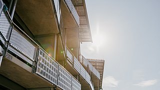 Blick von unten auf Balkone eines modernen Gebäudes mit Metallgeländern und Sonnenschutz, Sonne im Hintergrund.