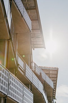 Foto, Blick von unten auf Balkone eines modernen Gebäudes mit Metallgeländern und Sonnenschutz, Sonne im Hintergrund.