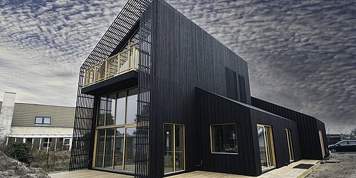 Foto, Modernes schwarzes Holzhaus mit einzigartiger schräger Architektur und großen Fenstern vor einem Wolkenhimmel.