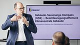 Foto, Mann im Anzug hält Mikrofon und spricht vor Publikum, Bildschirm zeigt Text zum Gebäude-Sanierungs-Kompass (GSK)