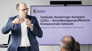 Foto, Mann im Anzug hält Mikrofon und spricht vor Publikum, Bildschirm zeigt Text zum Gebäude-Sanierungs-Kompass (GSK)