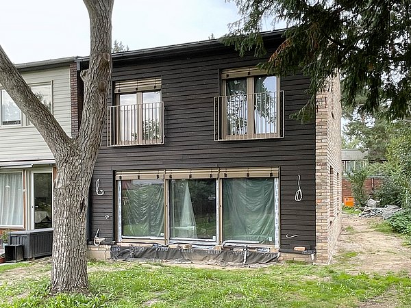 Foto, Zweistöckiges Neubau-Strohhaus mit dunkler Holzfassade, großen Fenstern und kleinem Balkon, umgeben von Gras und Bäumen